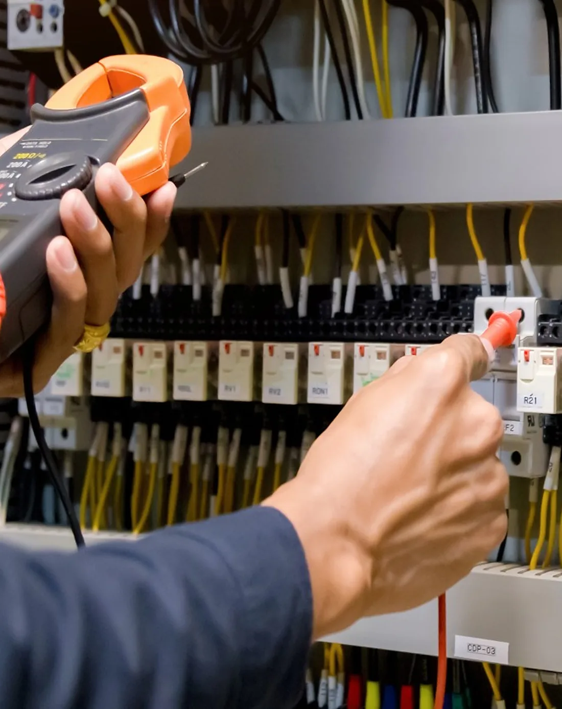 NEEC technician testing terminal blocks in a control panel with a clamp meter and test probe